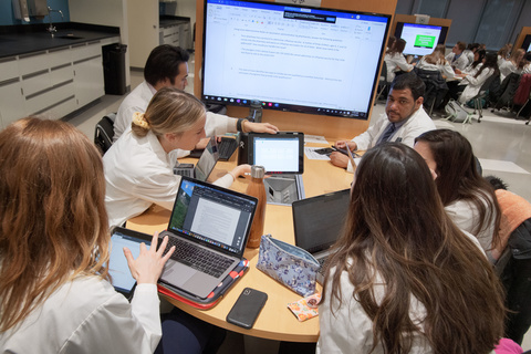 Students gather around screens in a large pharmacy practice classroom.