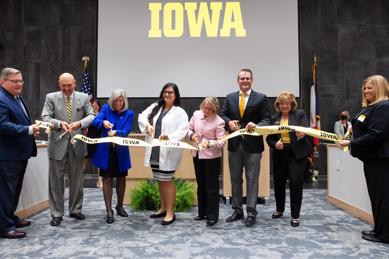 Six people cut a gold Iowa banner with large golden scissors