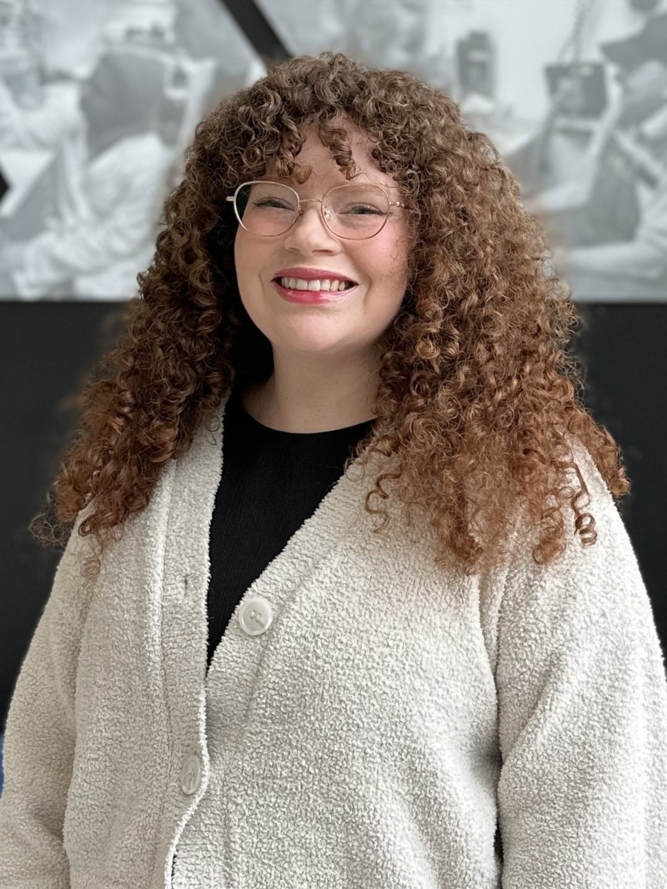 Portrait of Fabiola Gallardo Penchi smiling while wearing glasses, a black top, and a cream textured cardigan. She stands in front of a University of Iowa College of Pharmacy wall display featuring black-and-white images of students engaged in pharmacy education.
