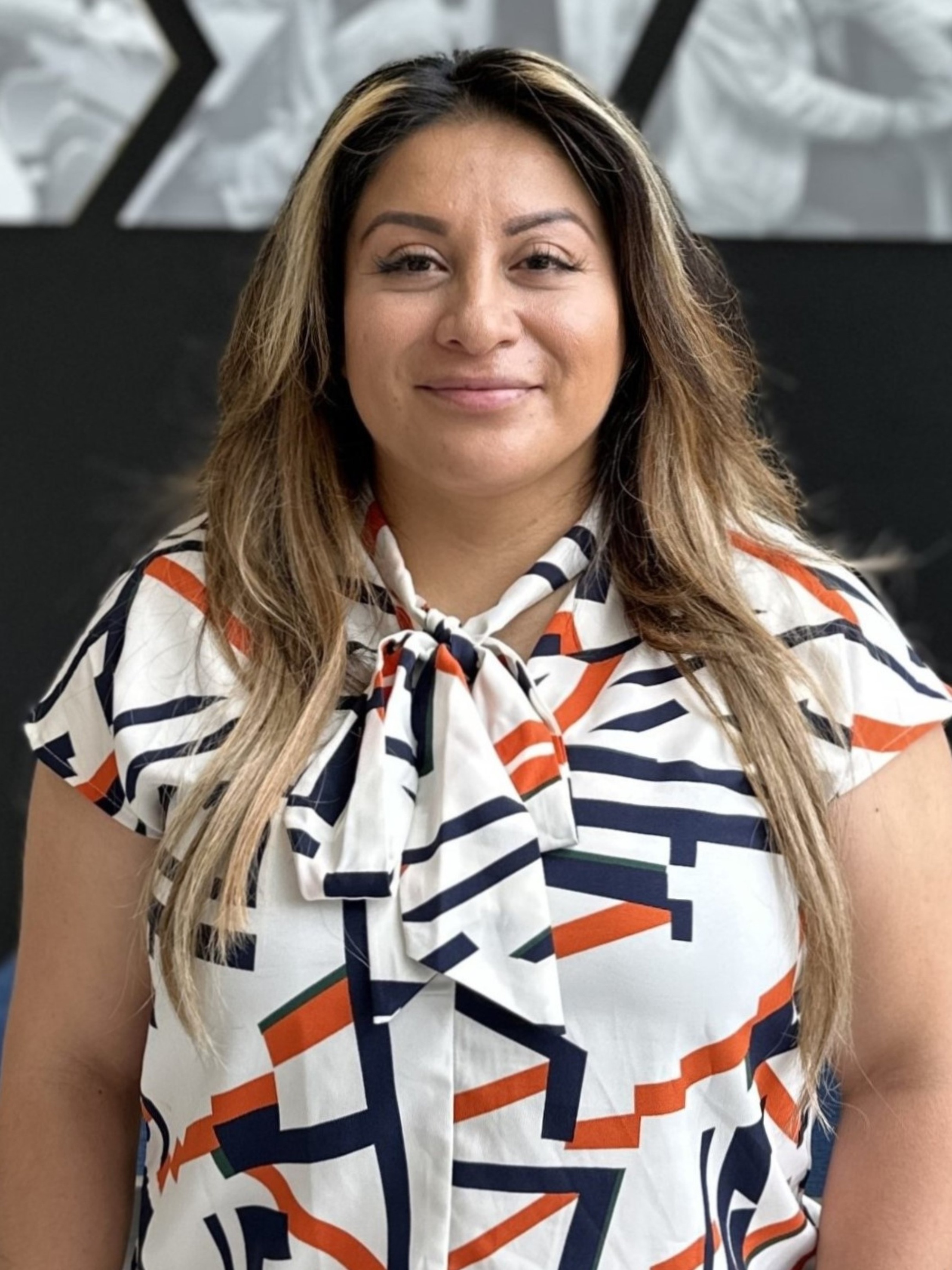 Portrait of Noemy Penafiel smiling at the camera while wearing a white blouse with a navy and orange geometric pattern and a tied neckline. She stands in front of a University of Iowa College of Pharmacy wall display featuring black-and-white images of students in learning and clinical environments.
