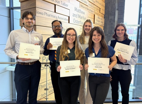 Group of six students stand together indoors, smiling and holding certificates recognizing research presentation awards. A donor recognition wall is visible behind them.