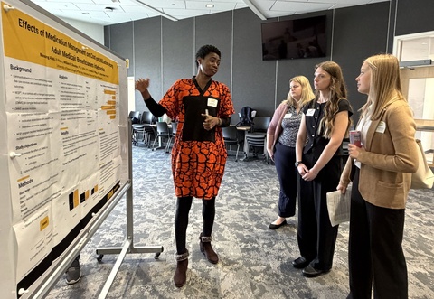 Student presents a research poster on medication management to three peers in a conference room. She gestures toward charts on the poster while explaining findings, as the others listen attentively. Chairs and a screen are visible in the background.