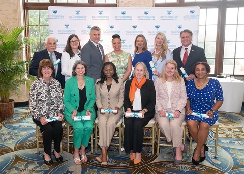 A group of well-dressed people posing in a group in front of a NACDS Foundation backdrop. There are people standing in the back and sitting in chairs in the front. All are holding a white piece of paper rolled and tied with a blue ribbon bow.