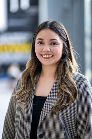Woman with wavy brown hair with lighter ends. She is wearing a black and white patterned jacket and a black top. She is standing in the College of Pharmacy with the background blurred.