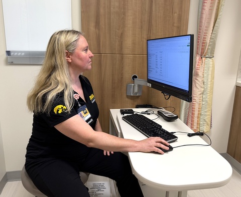 A pharmacist sits at a computer workstation in a clinic exam room, using a mouse while viewing a patient care spreadsheet on a monitor. She wears a black University of Iowa Pharmacy polo shirt with an ID badge. The room includes a keyboard, scanner, wood cabinetry, privacy curtain, and an exam table partially visible in the background.
