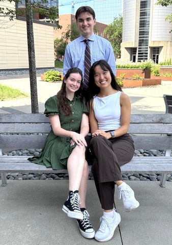Three University of Iowa College of Pharmacy students pose on a bench outside the College of Pharmacy building. Two students sit on the bench while a third stands behind them, smiling with campus buildings and landscaping in the background.