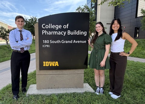 Three University of Iowa College of Pharmacy students pose beside the College of Pharmacy building sign reading "180 South Grand Avenue." They smile outdoors on a sunny day with the building and surrounding campus visible behind them.