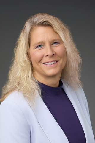 Professional headshot of Laura Borgelt, a smiling woman with long, wavy blonde hair. She wears a light-colored blazer over a dark purple high-neck top and faces the camera against a plain dark gray background.