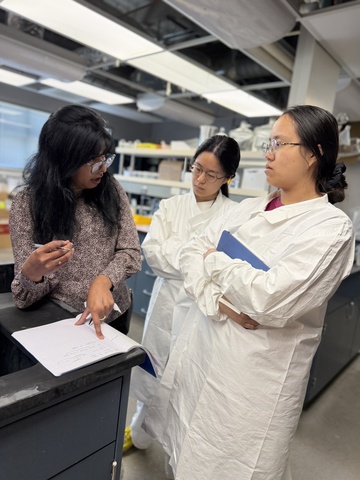 Bindu Gottam, University of Iowa Pharmaceuticals director of Preformulation and Formulation Development, stands at a black laboratory counter with a pen and notebook explaining to two students lyophilization process fundamentals and key considerations for successful product development. The two students wear white lab coats and are listening. Laboratory shelves and bright overhead lights fill the background.
