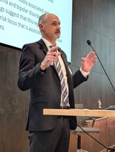 Speaker in a suit presents at a podium, gesturing with both hands while addressing an audience. A projected slide with text is visible behind him, and a microphone stands at the podium.