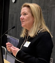Speaker stands at a podium, addressing an audience while gesturing with her hands. A microphone and computer monitor are in front of her in a lecture hall setting.