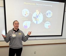 Pharmacist presents in a classroom beside a slide titled “Pharmacists: Beyond Dispensing,” explaining roles like medication optimization, patient education, chronic disease management, and adherence support while gesturing to the screen.