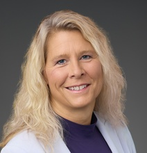 Smiling woman with long blonde hair wearing a light-colored blazer over a purple top, photographed against a dark gray studio background.