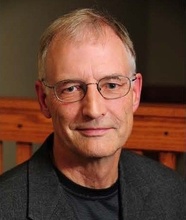 Man with short gray hair and glasses wearing a dark jacket over a black shirt, photographed indoors with a wooden railing in the background.