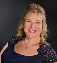 miling woman with blonde hair wearing a navy dress with lace detailing and a necklace, photographed against a dark studio background.