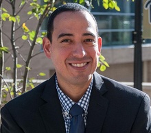 Smiling man with short dark hair wearing a dark suit, blue-and-white checkered shirt, and blue tie, with an indoor background featuring a plant.