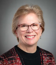 Smiling woman with short blonde hair and glasses wearing a red, black, and white patterned jacket over a black top against a gray background.