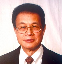 Man with short dark hair and glasses wearing a dark suit, white shirt, and patterned tie, photographed against a light-colored background.