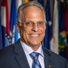 Smiling man with short gray hair and glasses wearing a dark suit, light blue shirt, and striped blue and purple tie, posed in front of a row of flags.