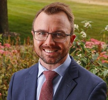 Smiling man with short brown hair, beard, and glasses wearing a navy suit, light blue shirt, and red tie, posed outdoors with flowers and greenery behind him.