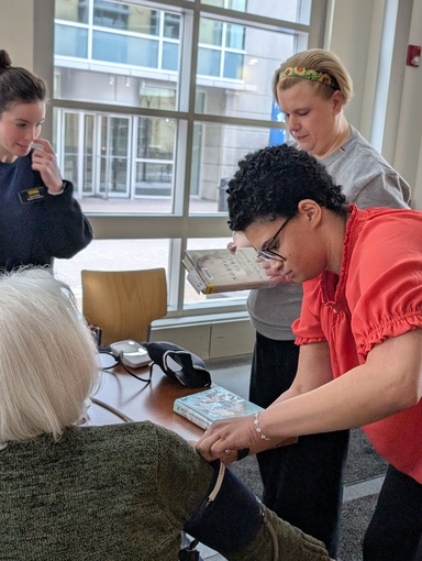 Two University of Iowa student pharmacists conduct a blood pressure screening at a library. One student in a red top leans forward to place a blood pressure cuff on a seated patron.with white hair, while another student observes from behind and a faculty member looks on.