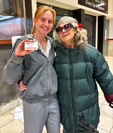 A pharmacy student in gray pants and a zip-up hoodie holds up a small card reading "I WILL TAKE MY MEDS." while posing with an elderly woman in a green puffer coat and large sunglasses at an indoor mall event.