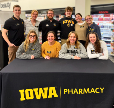 Ten University of Iowa Pharmacy students pose for a group photo at a CVS store, with four seated in front and six standing behind them. They are gathered around a table draped with a black tablecloth printed with the Iowa Pharmacy logo in gold.