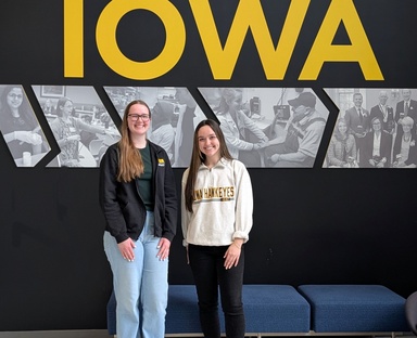 Two young women standing in front of a black wall that reads "IOWA" in gold letters. On the left: a tall woman with glasses in a black Iowa Pharmacy zip-up. On the right: a shorter woman in a white Iowa Hawkeyes zip-up. Black and white students of faculty are behind them on the wall.