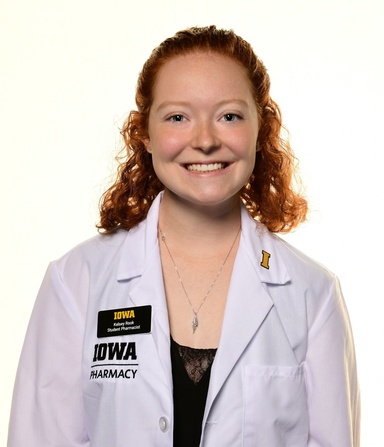 Smiling student pharmacist with curly red hair wears a white Iowa Pharmacy coat with a name badge reading “Kelsey Rook, Student Pharmacist” and a gold Iowa “I” pin against a plain white background.