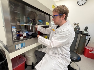 Researcher Dane Lew, a member of University of Iowa College of Pharmacy Professor Michael Ashley Spies’ lab, is wearing a white lab coat, safety glasses, and gloves using a pipette while working inside a laboratory biosafety cabinet.