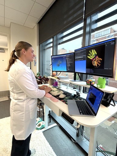 University of Iowa College of Pharmacy PhD student and researcher Lilly Duff wears a white lab coat while working at a computer station displaying colorful molecular structures and computational modeling data on two large monitors. She is part of University of Iowa College of Pharmacy Professor Michael Ashley Spies’ lab.