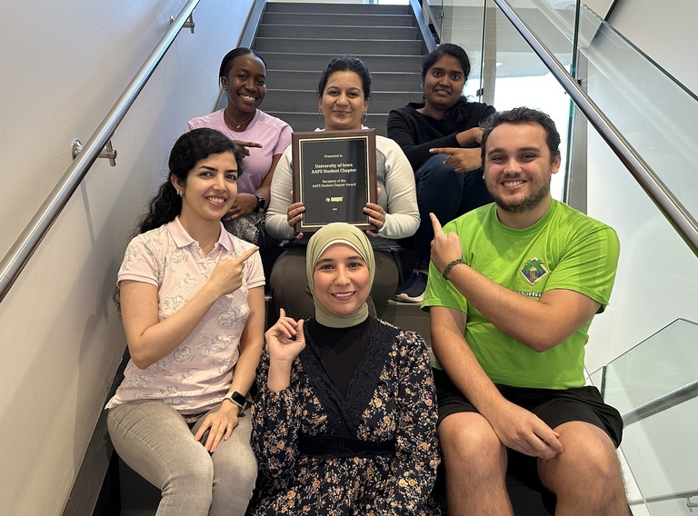 Six students sit on stairs with the middle person holding a chapter award plaque and the  others pointing at her and the plalque.