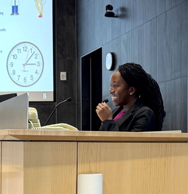 A student smiles while speaking into a microphone at a podium in a lecture hall. A presentation slide with graphics and a clock is visible behind her as she delivers her talk.