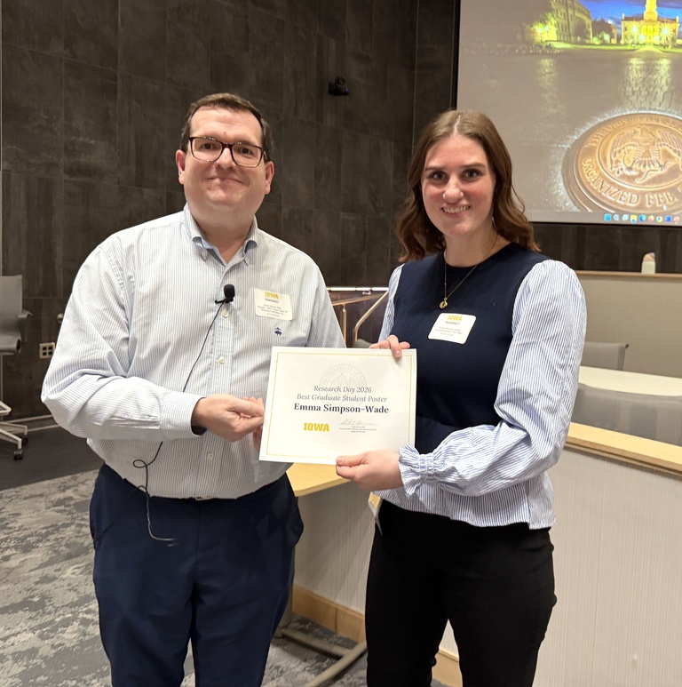 Two people stand together holding a certificate that reads “Research Day 2026 Best Graduate Student Poster.” Both wear name tags and smile at the camera in a lecture hall setting with a presentation screen in the background.