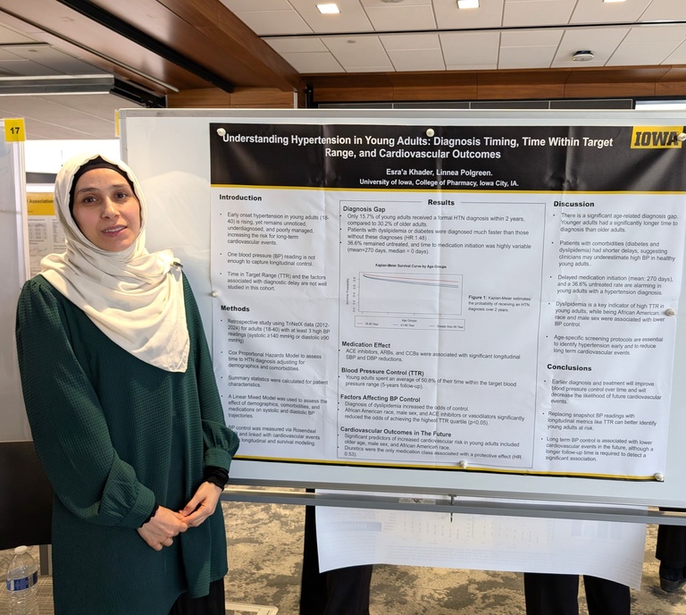 A woman stands beside a research poster titled “Understanding Hypertension in Young Adults,” presenting findings on diagnosis timing and cardiovascular outcomes. She faces the camera in a conference setting with poster boards and numbered displays in the background.