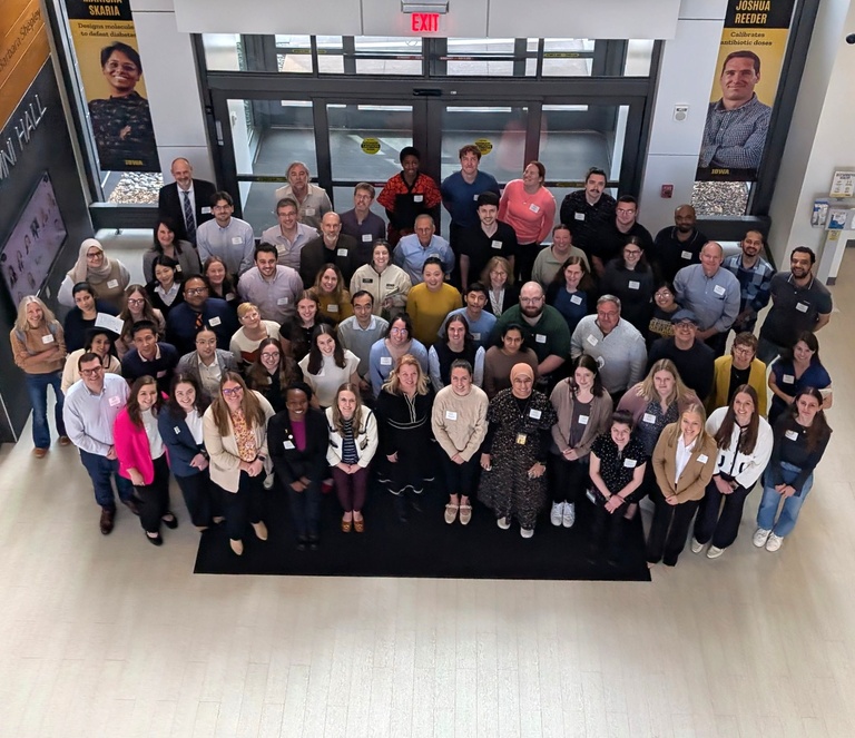 A large group of students, faculty, and staff stand together in a building lobby, smiling up at the camera for a group photo. They wear name tags and are gathered near glass entrance doors, with campus banners visible on the walls.