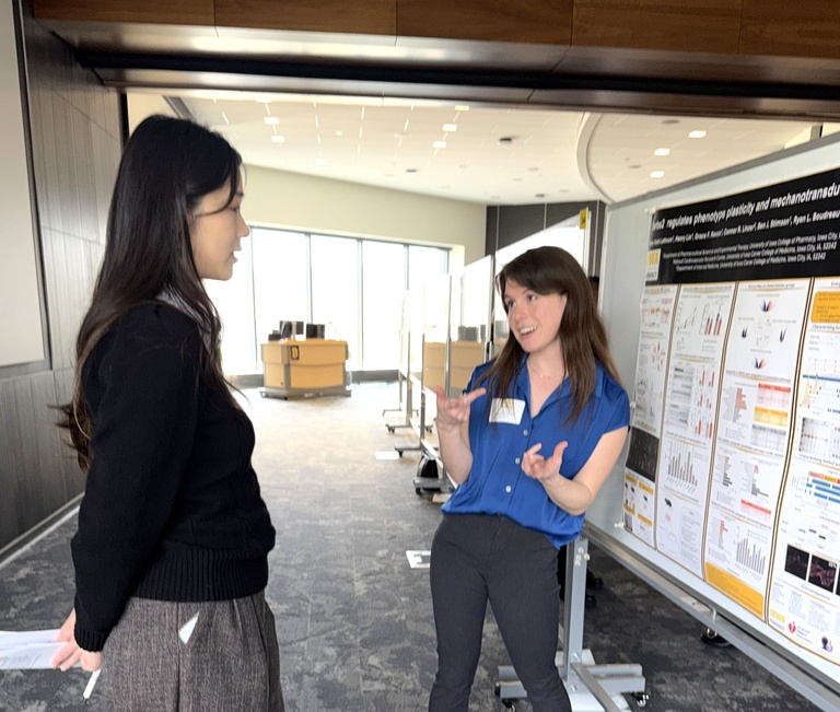 Two women stand in a bright indoor space beside a scientific research poster. One gestures while explaining the poster’s charts and data, and the other listens attentively, holding notes. Portable poster boards and large windows are visible in the background.