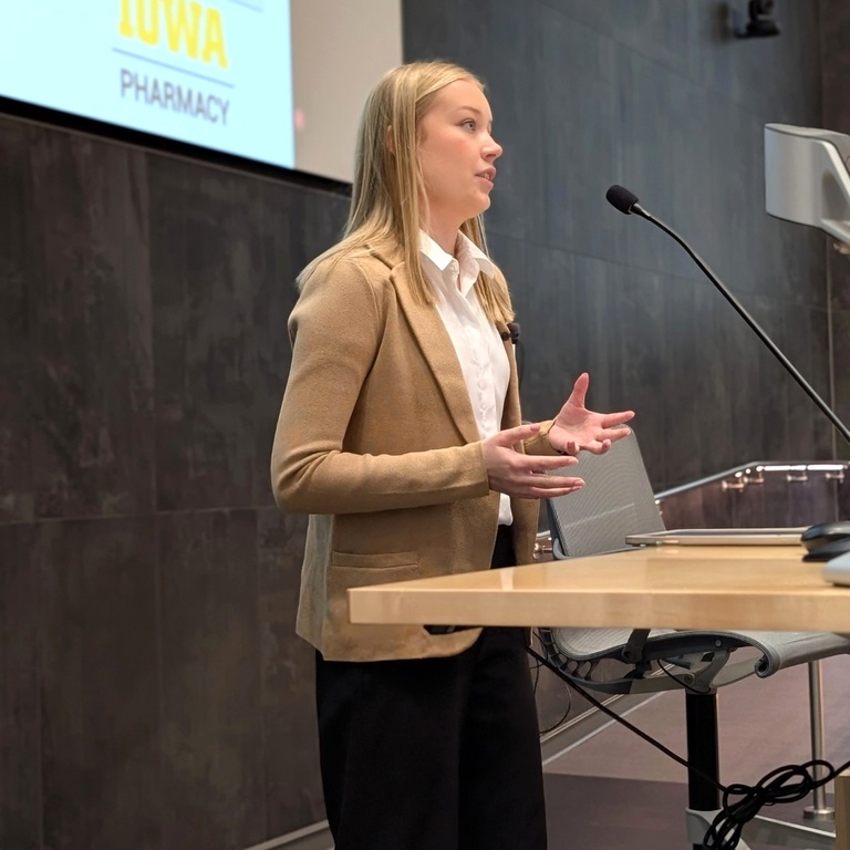 A student stands at a podium giving a presentation, speaking into a microphone while gesturing with her hands. A screen displaying “Iowa Pharmacy” is visible behind her in a lecture hall setting.