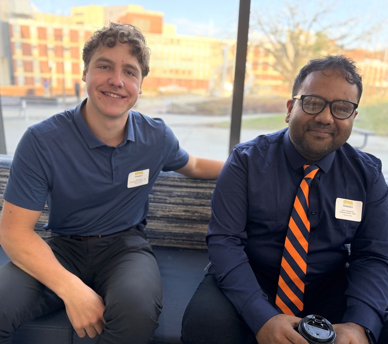 Two men sit side by side on a bench indoors, smiling at the camera. Both wear name tags; one is dressed in a blue polo, and the other in a dark shirt with an orange striped tie, holding a coffee cup. Large windows behind them show a campus building outside.1
