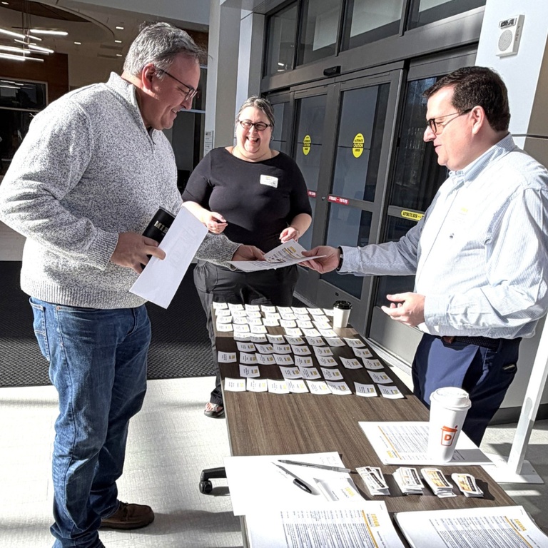 Three people stand at a registration table in a building lobby, handing out name tags and materials for an event. One person reaches to receive a badge while others assist, with rows of name tags, papers, and coffee cups arranged on the table.