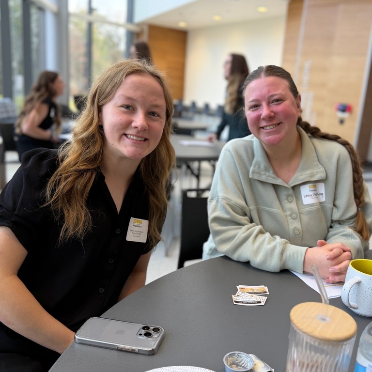 Two students sit at a table indoors, smiling at the camera during a campus event. Both wear name tags and have drinks and small items on the table, with other attendees and large windows visible in the background.