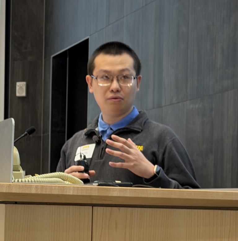 Presenter stands at a podium speaking to an audience, gesturing with one hand while holding a clicker. A slide with text is projected behind him, and a computer monitor sits on the podium.