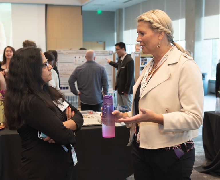 Two women talk face-to-face at an indoor event. The woman on the left has long dark hair and glasses and wears black with her arms crossed. The blonde woman on the right, named Laura Borgelt, wears a light-colored jacket and conference lanyard, holds a purple-and-pink water bottle, and gestures while speaking. Several people and research poster displays are visible in the background.