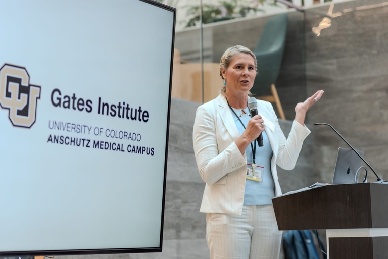 Laura Borgelt, a woman in a white pinstriped suit and light blue top, speaks into a handheld microphone at a podium, raising one hand as she addresses an audience. A laptop sits on the podium. Beside her, a large screen displays the CU logo and the words “Gates Institute, University of Colorado Anschutz Medical Campus.”