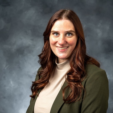 Photo of a white woman smiling against a gray background. Her body is turned to the left. The photo is taken from chest upwards. She has long, wavy red hair and blue eyes. She is wearing a white turtleneck and green blazer.