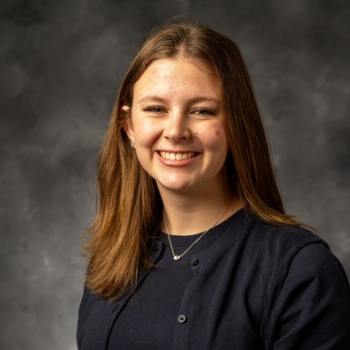 Photo of a white woman smiling at the camera against a gray background. She has straight, light brown hair to her shoulders. She is wearing a blue shirt and cardigan.