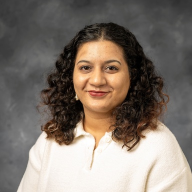 Photo of an Indian woman smiling against a gray background. Headshot is cropped from chest up. She has curly black hair with reddish-brown ends, and it falls to her shoulders. She is wearing a white button-up.