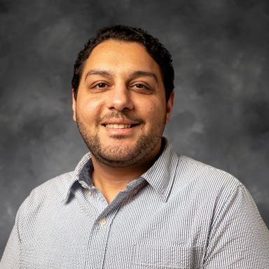 Photo of a middle-eastern man smiling against a gray background. Photo is taken from the chest-up. He has short black hair and facial hair. He is wearing a blue patterned button-up.