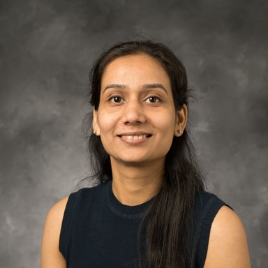 Headshot from the chest upwards. An Indian woman smiling at the camera against a gray background. She has long, straight black hair in a half-up half-down hairstyle. She is wearing a blue sleeveless tank.