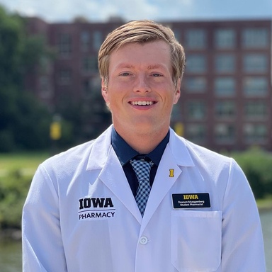 Dawson Mueggenberg smiles outdoors while wearing a white Iowa Pharmacy coat, dress shirt, and patterned tie. A campus building and water feature are blurred in the background on a sunny day.
